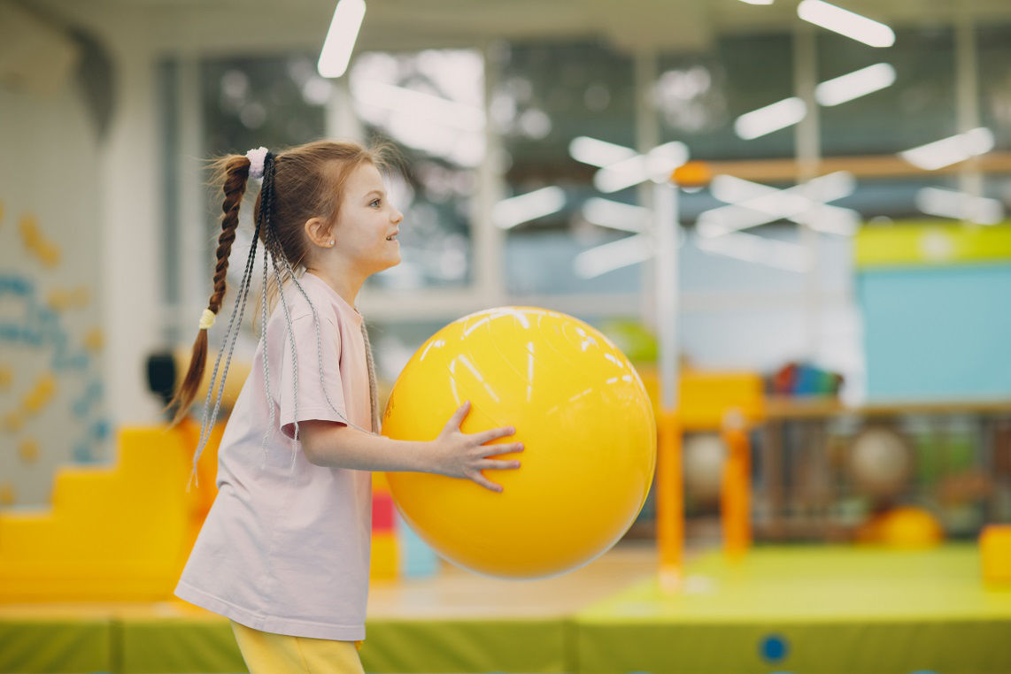 Little girl playing and doing exercises with big yellow ball in gym at kindergarten or elementary school. Children sport and fitness concept